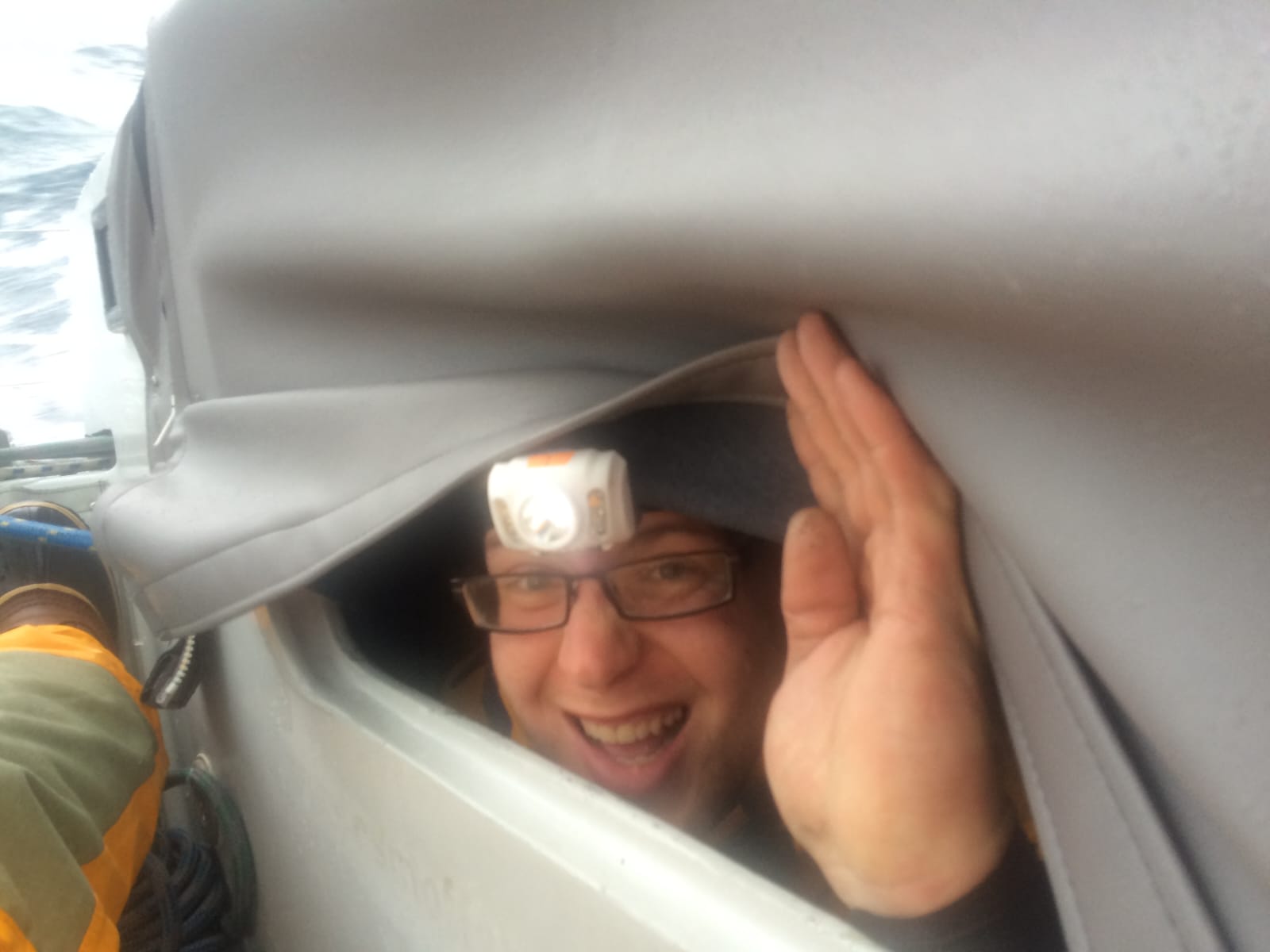 Ken peeking out from under a fabric cover during the storm, grinning, wearing a headlamp, boat heeled over